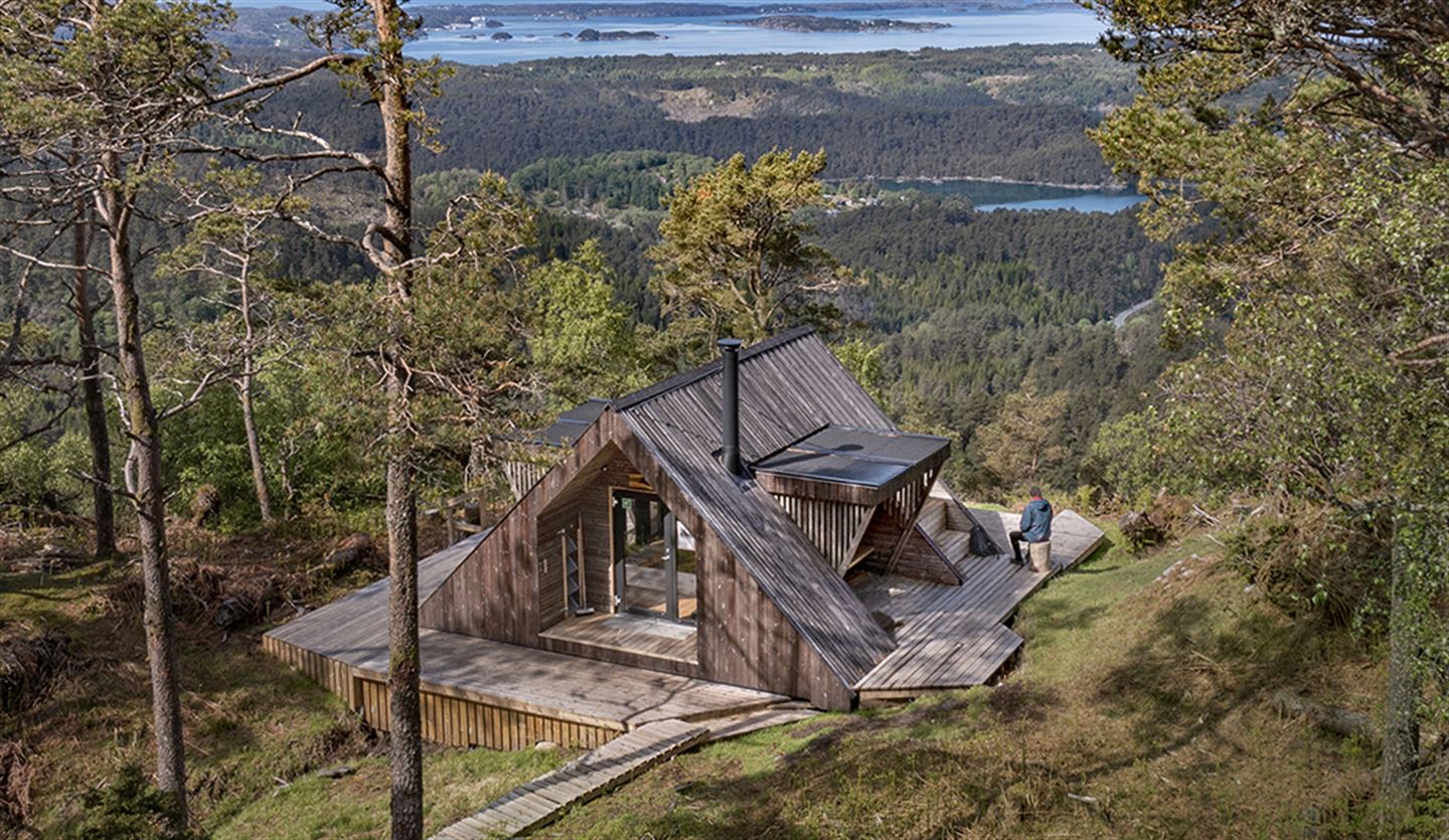 Arkitekttegnet dagsturhytte på en fjellside med utsikt over fjorden, omgitt av furutrær